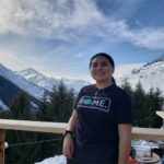 Nizhoni Tallas has dark hair pulled back in a ponytail and wears a navy blue teeshirt that reads "home" She stands in front of a landscape of snowy mountains.