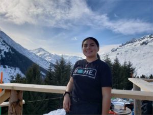 Nizhoni Tallas has dark hair pulled back in a ponytail and wears a navy blue teeshirt that reads "home" She stands in front of a landscape of snowy mountains.